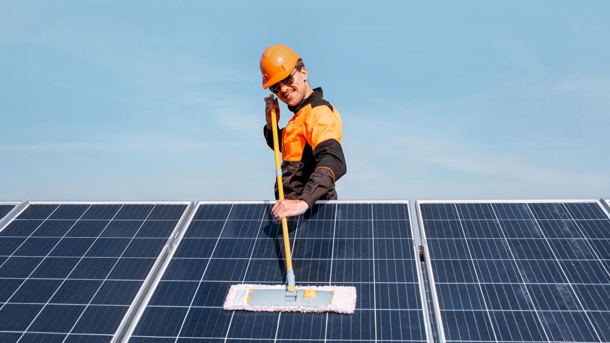 A worker in an orange safety helmet and orange and black uniform cleaning solar panels with a long handle brush against a clear blue sky.